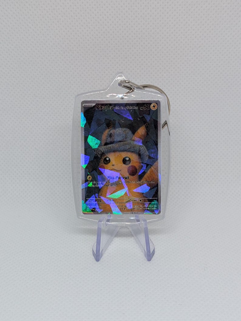 May include: A clear acrylic keychain containing a holographic trading card featuring a yellow Pikachu wearing a grey hat. The card displays the text Pikachu, Pika-Portrait, and 60 HP. The keychain has a metal ring at the top.