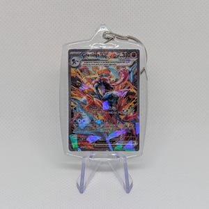 May include: A clear acrylic keychain case containing a Greninja ex trading card with 310 HP. The card features a holographic illustration of the blue and red character with the text Shinobi Blade and Mirage Barrage printed on the front.
