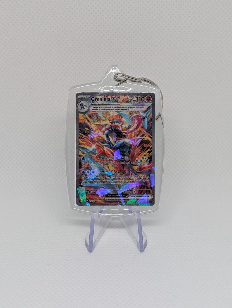 May include: A clear acrylic keychain case containing a Greninja ex trading card with 310 HP. The card features a holographic illustration of the blue and red character with the text Shinobi Blade and Mirage Barrage printed on the front.