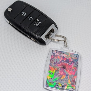 May include: A black car key fob with three buttons attached to a silver metal keyring holding a rectangular clear plastic case containing a Gengar VMAX trading card with a holographic finish and visible text including Gengar VMAX.
