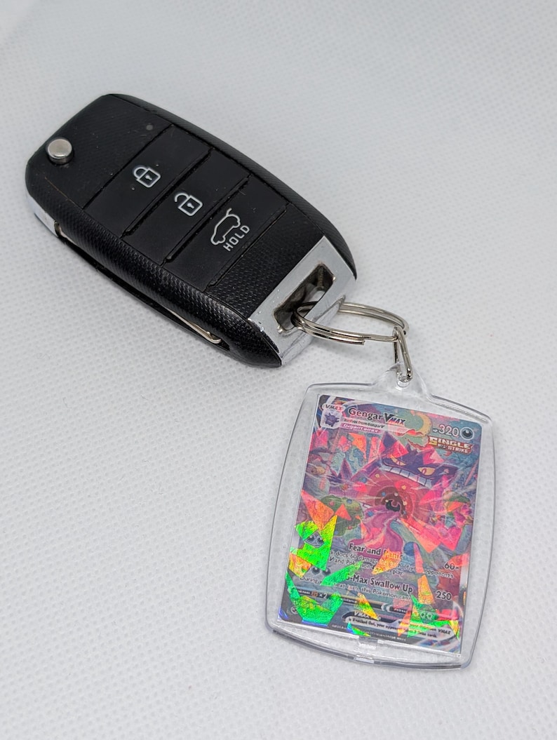 May include: A black car key fob with three buttons attached to a silver metal keyring holding a rectangular clear plastic case containing a Gengar VMAX trading card with a holographic finish and visible text including Gengar VMAX.