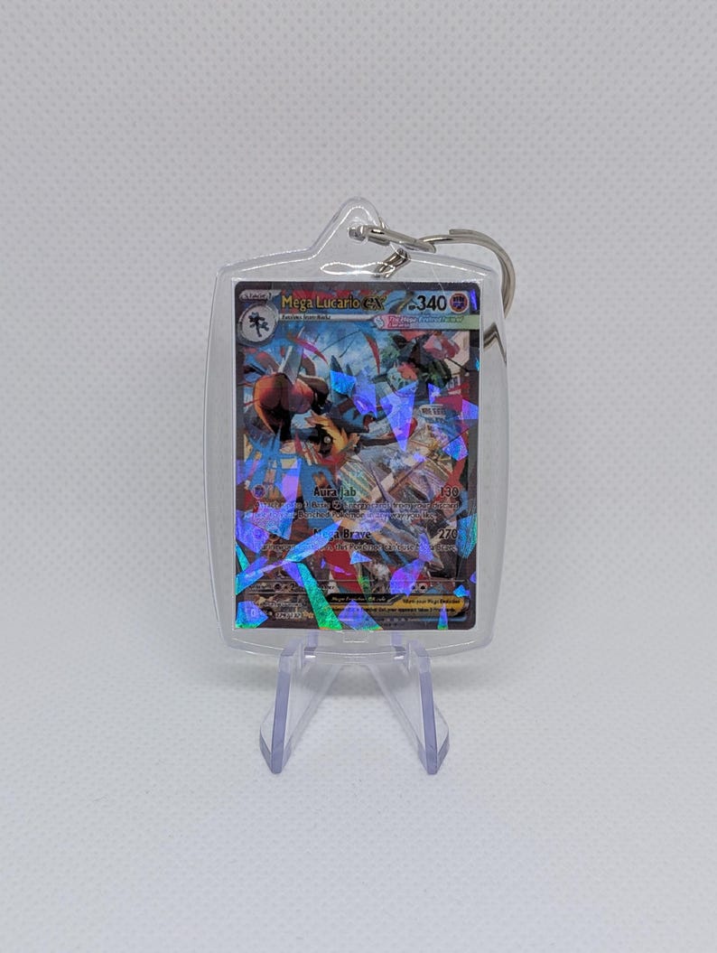 May include: A clear acrylic keychain featuring a Mega Lucario EX trading card with a holographic shattered glass finish. The card displays the character Lucario, the text Mega Lucario EX, and a health value of 340. A metal ring is attached to the top.