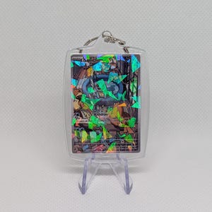 May include: A clear acrylic keychain case containing a Squirtle trading card with a holographic shattered glass finish. The card features the character in a blue pool, with text including Withdraw and Skull Bash visible through the plastic.