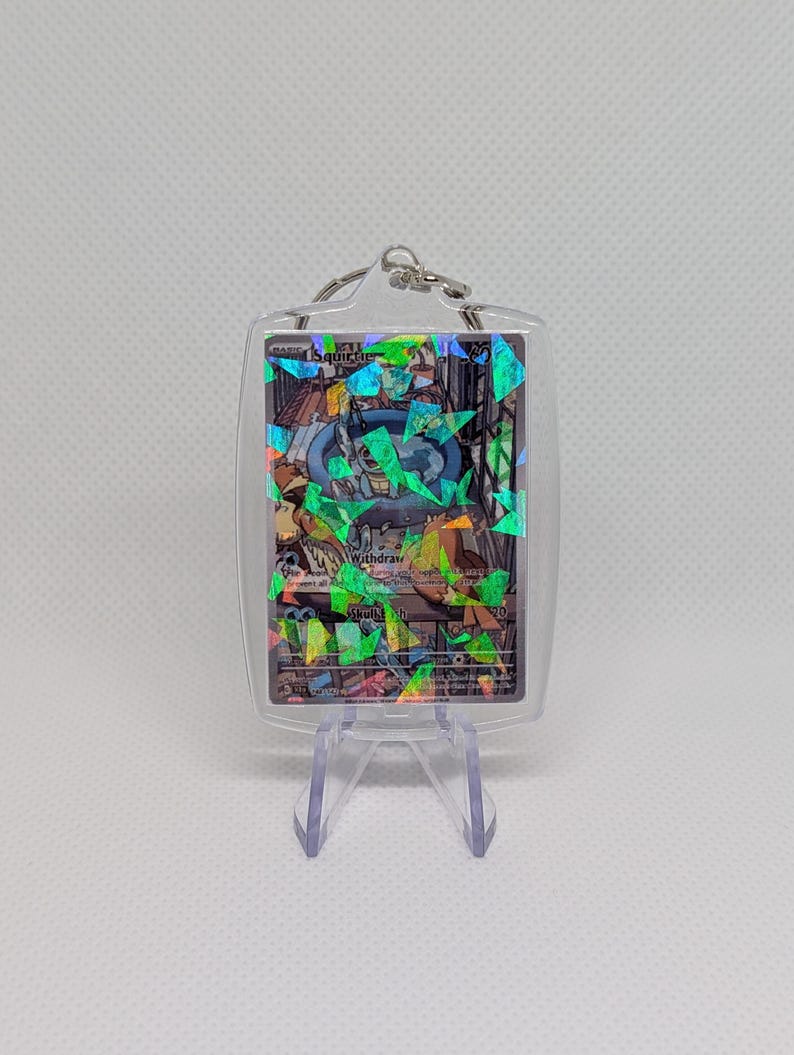 May include: A clear acrylic keychain case containing a Squirtle trading card with a holographic shattered glass finish. The card features the character in a blue pool, with text including Withdraw and Skull Bash visible through the plastic.
