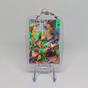 May include: A clear acrylic keychain featuring a holographic trading card of the character Bulbasaur. The card displays the name Bulbasaur, 70 HP, and the move Leech Seed. The holographic finish creates a shattered glass effect over the illustration of the character in a room setting. The keychain is held in a transparent protective case with a metal ring at the top.