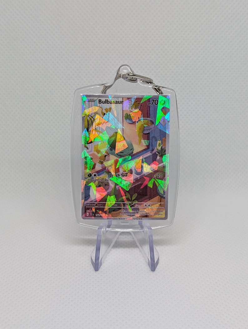 May include: A clear acrylic keychain featuring a holographic trading card of the character Bulbasaur. The card displays the name Bulbasaur, 70 HP, and the move Leech Seed. The holographic finish creates a shattered glass effect over the illustration of the character in a room setting. The keychain is held in a transparent protective case with a metal ring at the top.