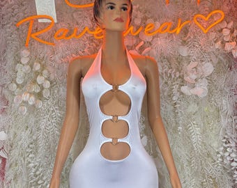 May include: White halter dress with three cut-out sections and gold-colored ring accents. The dress is displayed on a mannequin. The background features a neon sign that reads "Rave Wear".