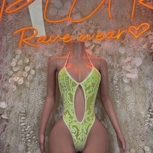 May include: Neon green bodysuit with a halter neck and a keyhole cutout. The bodysuit features a white trim and a decorative pattern. The background includes the neon text "Rave wear".