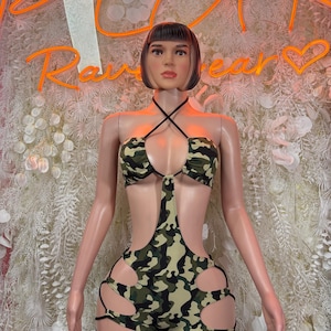 May include: A mannequin displaying a two-piece camouflage outfit. The top is a halter-neck bikini, and the bottoms are shorts with cut-out details and black ties. The background includes a neon sign that reads "Raw Wear".
