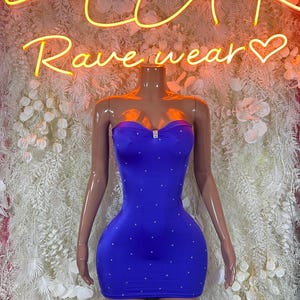May include: A royal blue mini dress with a strapless design, embellished with small, sparkling details. The dress, with a sweetheart neckline, is displayed on a mannequin. A neon sign in the background reads "PLUR Rave wear".