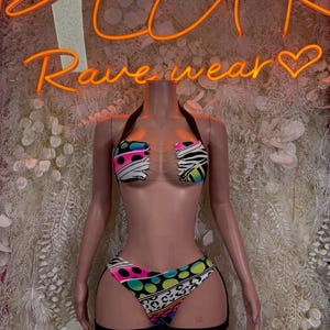 May include: A two-piece bikini set featuring a halter-style top and high-cut bottoms with a vibrant multi-colored pattern of neon polka dots, zebra stripes, and leopard spots, connected by clear plastic straps at the center of the chest.