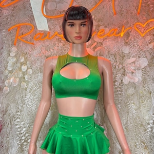 May include: A green two-piece outfit on a mannequin. The top has a keyhole cutout and the skirt is a ruffled mini skirt. The outfit is embellished with small, sparkling accents. The background features a neon sign that says "Rain Wear".
