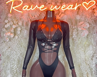 May include: Black bodysuit with a halter top and long sleeves, embellished with rhinestones. The text "PLUR Rave wear" is displayed in neon lights above the mannequin. The background is a white floral arrangement.