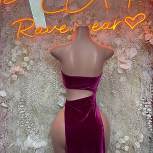 May include: A deep magenta velvet dress featuring a strapless bodice, a side cutout at the waist, and a high-thigh slit. The garment is displayed on a mannequin, showcasing the smooth texture of the fabric and the fitted silhouette of the design.