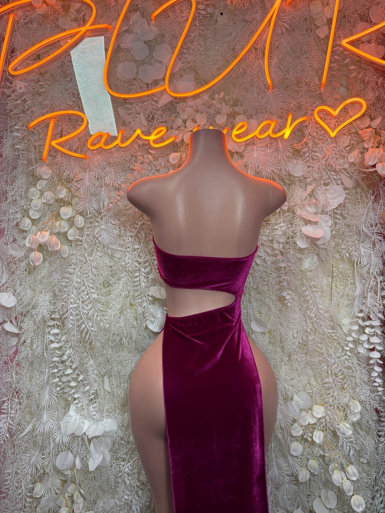 May include: A deep magenta velvet dress featuring a strapless bodice, a side cutout at the waist, and a high-thigh slit. The garment is displayed on a mannequin, showcasing the smooth texture of the fabric and the fitted silhouette of the design.