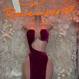 May include: A deep magenta velvet dress featuring a strapless bodice with a lace-up front detail, a large side cutout, and a high-thigh slit. The garment is displayed on a mannequin, showcasing the soft texture of the fabric and the fitted silhouette.