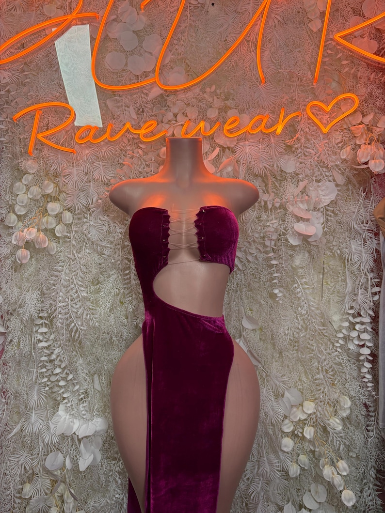 May include: A deep magenta velvet dress featuring a strapless bodice with a lace-up front detail, a large side cutout, and a high-thigh slit. The garment is displayed on a mannequin, showcasing the soft texture of the fabric and the fitted silhouette.