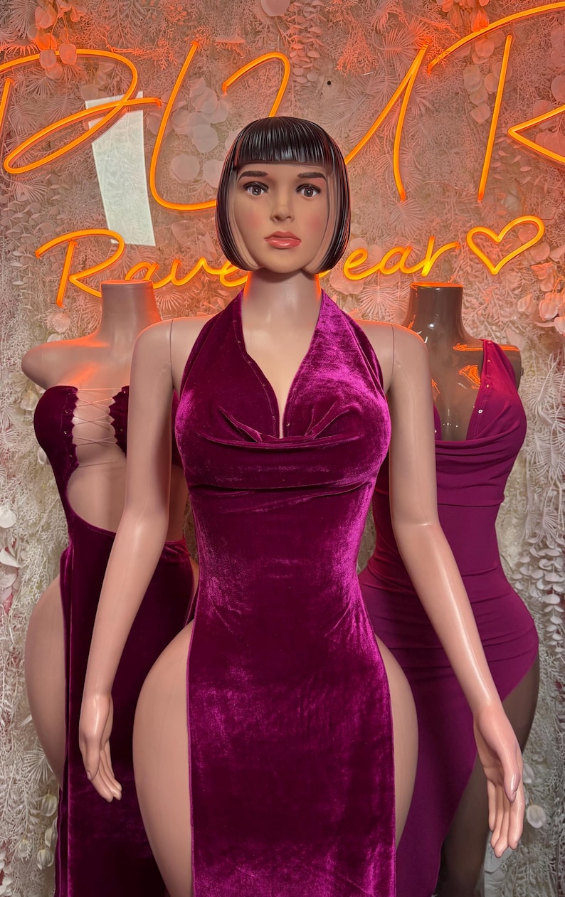 May include: A dark magenta velvet halter dress with a cowl neckline and high side slits, displayed on a mannequin with a short black bob hairstyle. The dress features a soft, plush texture and a fitted silhouette that extends to the floor.
