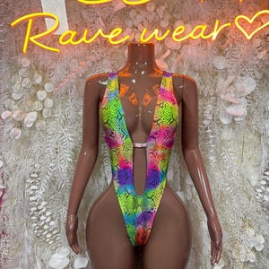 May include: A sleeveless one-piece bodysuit featuring a vibrant, multi-colored snakeskin pattern in neon yellow, pink, blue, and green. The garment has a deep, plunging V-neckline that extends to the waist, connected by a small horizontal strap.