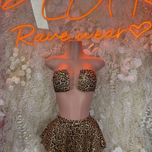 May include: A leopard print two-piece rave outfit. The set includes a strapless top and a ruffled mini skirt. The outfit is displayed on a mannequin against a backdrop with the neon text "Rave wear".
