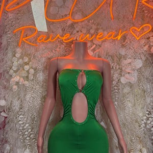 May include: A green strapless mini dress with a keyhole cutout and gold-colored accents. The dress is displayed on a headless mannequin. The background features a neon sign that reads "PLUR Rave wear".