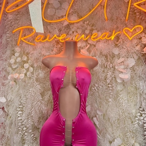 May include: A hot pink, strapless mini dress with a cut-out design. The dress features a series of small, decorative buttons and delicate chain accents. The background includes a neon sign that reads "PLUR Ravewear".