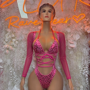 May include: A pink and black leopard print bodysuit featuring a deep V-neckline, thin shoulder straps, and a lace-up front detail. The garment includes long, sheer pink mesh sleeves and a high-cut leg design, displayed on a mannequin.