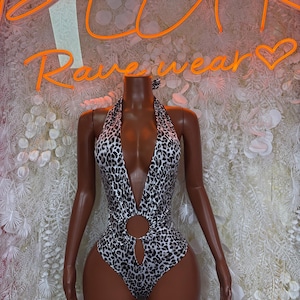 May include: A sleeveless one-piece bodysuit featuring a black and white leopard print pattern. The garment has a deep plunging halter neckline and a circular cutout detail at the center of the torso, creating a high-cut leg silhouette.