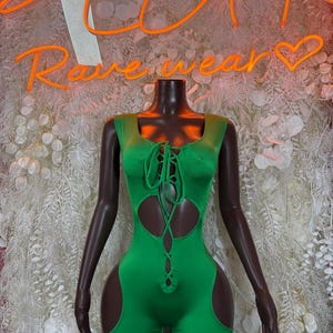 May include: A vibrant green romper with a lace-up front and cut-out details. The sleeveless garment is displayed on a mannequin. The neon sign behind the mannequin reads "Rave Wear".
