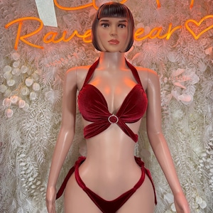 May include: A red velvet lingerie set on a mannequin. The halter top features a central ring detail, and the bottoms have tie closures. The background includes a floral wall and neon sign that reads "Rave Wear".
