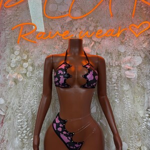 May include: A black and pink patterned bikini set on a mannequin. The bikini top and high-waisted bottoms feature a cow print design with clear straps. The neon sign behind the mannequin reads "Rave Wear".