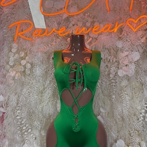 May include: A bright green bodysuit with cut-out details and a lace-up front. The sleeveless garment features a scoop neckline and short shorts. The neon sign in the background reads "Rave wear".
