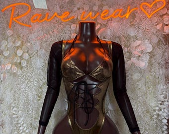May include: Gold metallic bodysuit with a halter neck and lace-up detail. The bodysuit has long, sheer black sleeves. The background features a neon sign that says "Rave Wear".