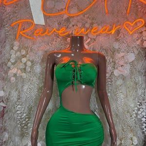 May include: A vibrant green mini dress with a strapless, lace-up bodice and a side cutout. The dress is displayed on a mannequin. The background features a neon sign that reads "Rave wear".