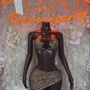 Peut inclure: Un mannequin présente un ensemble deux pièces de rave à imprimé léopard. L'ensemble comprend un haut dos nu avec des détails croisés et une mini-jupe asymétrique assortie. Le fond présente un texte orange néon qui se lit "PLUR Rave wear".