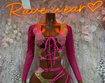 May include: A two-piece rave outfit with a vibrant, swirling pattern in pink, green, and yellow. The top features long pink mesh sleeves and a lace-up front. The matching shorts have cut-out details and pink straps. The neon sign behind the outfit says "Rave wear".