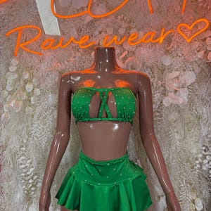 May include: A green two-piece rave outfit on a mannequin. The top is a strapless bandeau with a cut-out design and the skirt is a ruffled mini skirt. Both pieces are embellished with small, white rhinestones. The neon sign behind the mannequin says "Rave wear".