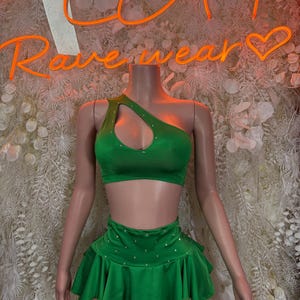 May include: A green two-piece outfit on a mannequin. The top is a one-shoulder design with a cutout and rhinestone embellishments. The matching skirt is a mini skirt with a ruffled hem and rhinestone accents. The neon sign behind the mannequin says "Rave wear".
