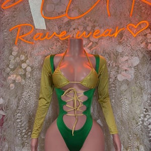 May include: A green and gold bodysuit with long sleeves and a halter top. The front of the bodysuit has a lace-up design. The words "Rave wear" are in neon orange above the mannequin.