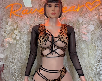May include: A two-piece leopard print outfit on a mannequin. The top has long black mesh sleeves and a lace-up front. The bottoms are high-waisted shorts with cut-out details. The background features a neon sign that says "Rave Wear".