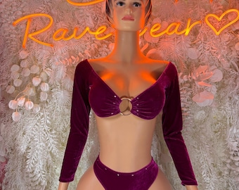 May include: A burgundy velvet bikini set with long sleeves. The top features a gold ring detail and the high-waisted bottoms have small embellishments. The background includes a neon sign that says "Rave Wear".