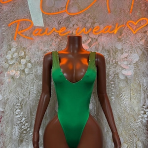 May include: A vibrant green bodysuit with a deep V-neck and rhinestone accents on the straps. The mannequin is positioned in front of a wall with the neon sign that reads "Rave wear".