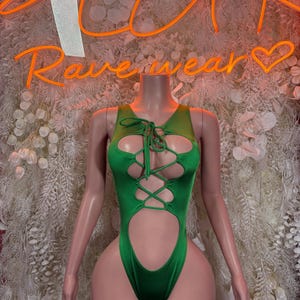 May include: A green bodysuit with a lace-up front and cut-out details. The neon sign behind the mannequin reads "Rave wear". The bodysuit is displayed on a mannequin with a neutral skin tone, set against a backdrop of white floral decorations.
