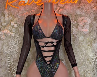 May include: A holographic bodysuit with a halter top and lace-up detail. The garment features long, sheer sleeves and is displayed on a mannequin. The background includes a neon sign that reads "Rave Wear".