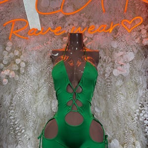 May include: A vibrant green halter-neck bodysuit with cut-out details and tie closures. The one-piece garment features a plunging neckline and shorts-style bottom. The background includes a neon sign that reads "Rave Wear".