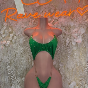 May include: A green bodysuit with gold-colored accents and a halter neck design. The bodysuit has a cut-out design and is embellished with small, gold-colored studs. The word "Rave wear" is written in orange neon lights in the background.
