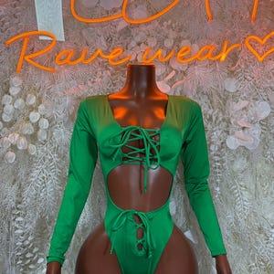 May include: A vibrant green bodysuit with long sleeves and a lace-up front and waist. The cutout design adds a modern touch to this stylish one-piece garment. The neon sign behind the bodysuit says "Ravewear".