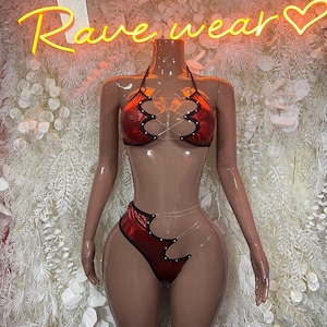 May include: A two-piece set featuring a red metallic halter top and matching high-cut bottom. Both pieces have black scalloped edges with small silver studs and are connected by thin, clear plastic straps that create a crisscross design.