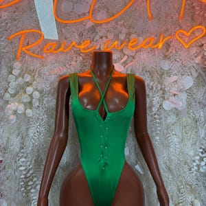 May include: A green bodysuit with a halter neckline and a high-cut leg. The bodysuit has a series of small, silver-colored buttons down the front. The words "Rave Wear" are displayed in neon orange in the background.
