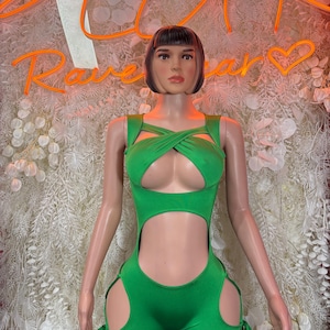 May include: A green romper with cut-out details and tie accents. The romper features a criss-cross design on the bodice and cut-outs on the sides of the shorts. The background includes neon orange text that reads "Rave Wear".